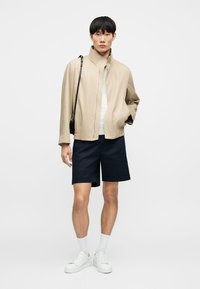 Jeune homme portant une veste beige, une chemise blanche, un short bleu marine, des chaussettes blanches, des baskets blanches, avec un sac à bandoulière noir sur un fond uni.