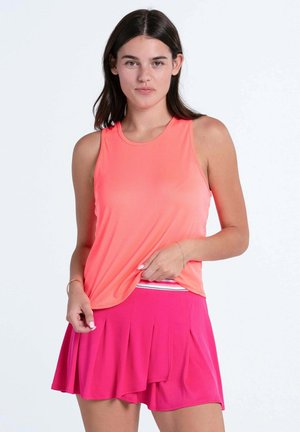 Frau trägt ein ärmelloses korallenfarbenes Oberteil und einen hellrosa plissierten Tennisrock, steht vor einem schlichten hellen Hintergrund.