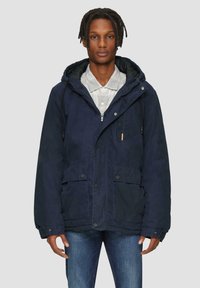 Marineblauwe hoodie met een ritssluiting, knoopzakken aan de voorkant en verstelbaar koord. Gepaarde met een lichtgrijs overhemd met kraag.