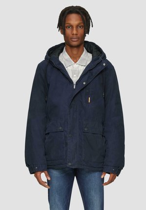 Veste à capuche bleu marine avec fermeture éclair, poches avant boutonnées et cordon de serrage réglable. Associée à une chemise à col gris clair.