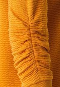 Texturovaný žlutý svetr s rukávem, který má nabíranou látku, s vroubkovaným manžetou a vlnitým vzorem po celé textilii pro přidanou dimenzi.