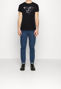 Sort bomulds T-shirt med et trekantet grafisk logo i hvid på brystet, parret med blå denimjeans og sorte ankelstøvler.