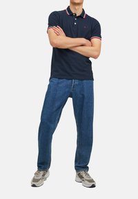 Marineblauwe polo met een rood-witte gestreepte kraag, gecombineerd met blauwe losvallende jeans en grijze sneakers. Het model poseert met de armen gekruist.