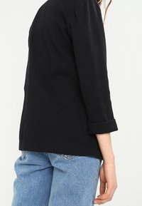 Černé tričko s dlouhým rukávem a ohrnutými manžetami, vyrobené z texturované látky. Má volnější střih a sahá pod pas, kombinováno s světle modrými džínami.