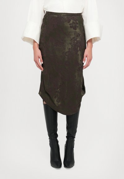 Satinrock in Olivgrün mit floralem Muster, asymmetrischem Saum und seitlichem Schlitz, kombiniert mit schwarzen knielangen, figurbetonten Stiefeln.