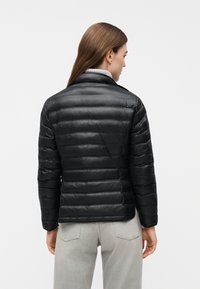 Femme vue de dos portant une veste matelassée noire et un pantalon gris clair, debout devant un fond blanc uni.