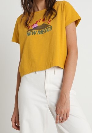 Nainen pukeutunut löysään keltaiseen lyhytmidikauluspaitaan, jossa on "New Mexico"-teksti ja grafiikka, yhdistetty korkeavyötäröisiin valkoisiin housuihin ja kahteen sormukseen oikeassa kädessä.