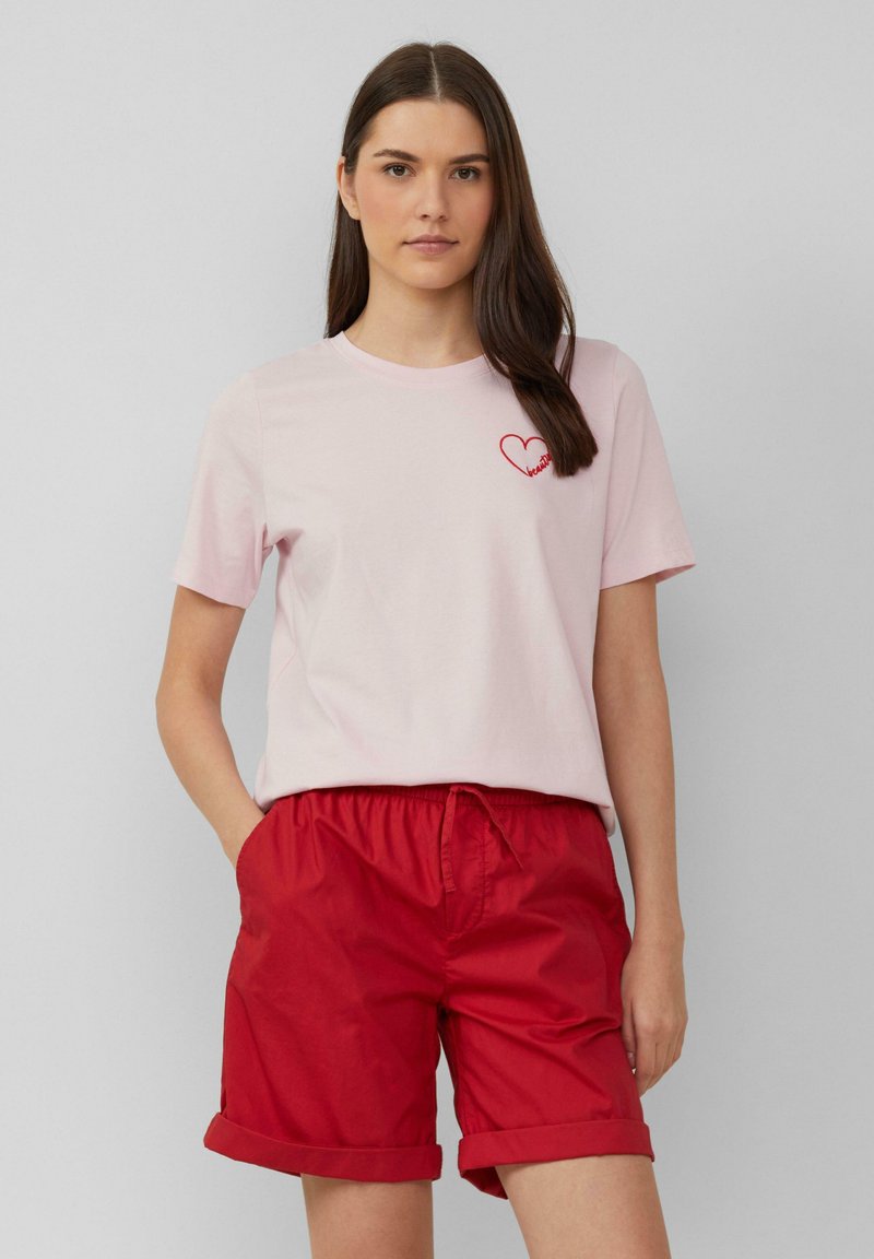 Lichtroze katoenen t-shirt met korte mouwen, met een rood hartlogo. Gecombineerd met rode shorts met elastische tailleband en omgeslagen mouwen.