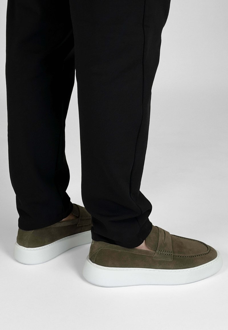 Olijfgroene suede instappers met witte rubberen zolen, met gestikte accenten en een gestructureerde afwerking, gecombineerd met zwarte broek.