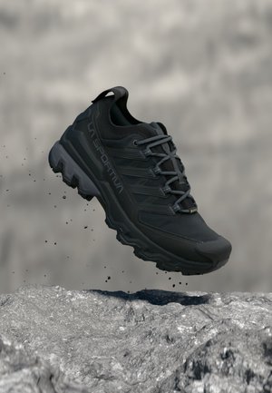 Chaussure de trail noire avec semelle rugueuse, marque La Sportiva et lacets gris, flottant au-dessus d'un terrain rocheux avec des particules de poussière.