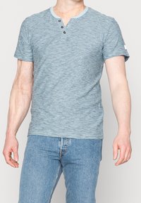 Modrá a bílá pruhovaná košile s krátkým rukávem, má výstřih Henley a tři knoflíky. Vyrobena z měkké, lehké tkaniny.