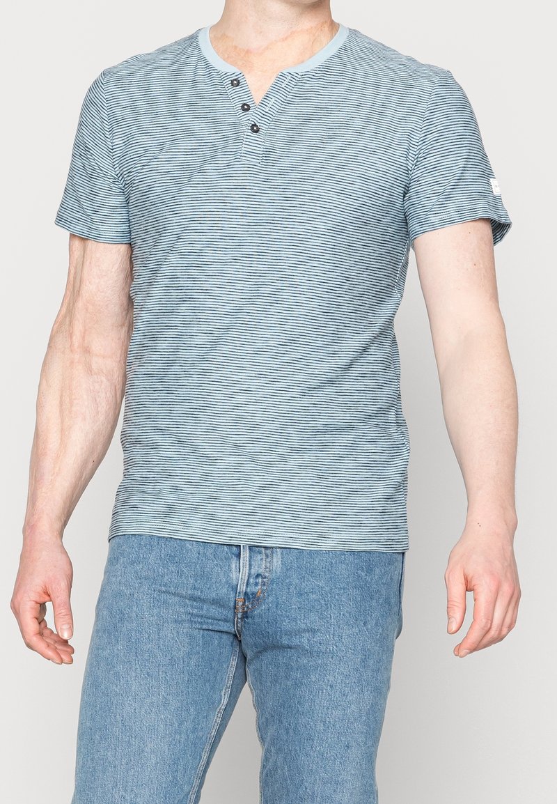 Modrá a bílá pruhovaná košile s krátkým rukávem, má výstřih Henley a tři knoflíky. Vyrobena z měkké, lehké tkaniny.