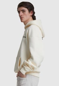 Kreemivärvi pullover-hoodie, millel on kängurupocket, ribitud mansetid ja tikitud must logo ees. Sile puuvillane kangas.