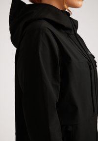 Giacca nera con cappuccio, caratterizzata da una superficie liscia, dotata di zip frontale, tasche laterali e cordini regolabili sul cappuccio.