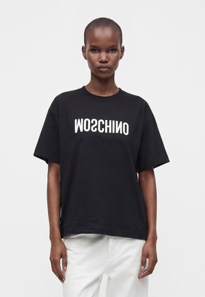 Μοντέλο με κοντά μαλλιά φοράει μαύρο φαρδύ t-shirt με λευκό ανεστραμμένο κείμενο "MOSCHINO" και λευκό παντελόνι σε απλό λευκό φόντο.