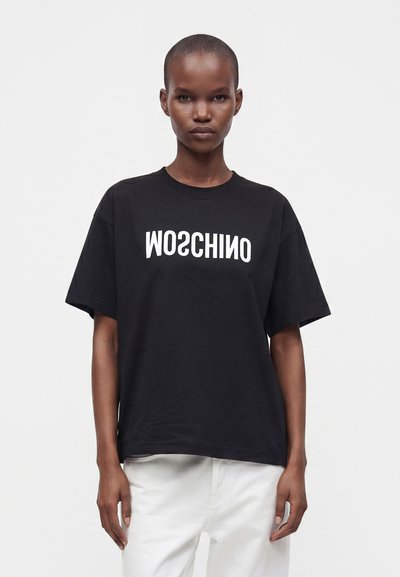 Μοντέλο με κοντά μαλλιά φοράει μαύρο φαρδύ t-shirt με λευκό ανεστραμμένο κείμενο "MOSCHINO" και λευκό παντελόνι σε απλό λευκό φόντο.