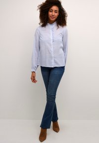 Blouse rayée bleu clair et blanche avec un col montant, un devant volé et des manches longues, associée à un jean en denim évasé et des bottes à talons marron.