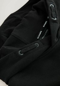 Černá bavlněná mikina s kapucí, vybavená šňůrkou s kulatými očky, žebrovanými manžetami a viditelnou štítkovou nášivkou na látce.