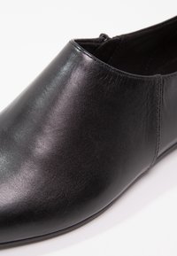 Chaussure noire en cuir à enfiler avec un bout pointu et une finition lisse, montrant un gros plan de la partie supérieure avant et d’un détail latéral.