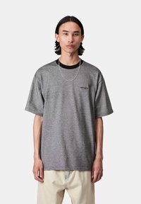 Młody mężczyzna z średniej długości czarnymi włosami, ubrany w szary t-shirt Carhartt, srebrny łańcuszek i jasnobeżowe spodnie na jednolitym tle.