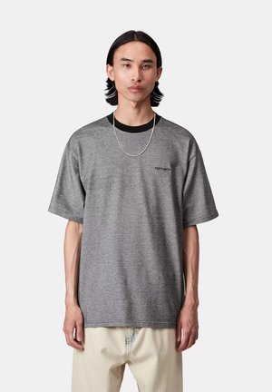 Młody mężczyzna z średniej długości czarnymi włosami, ubrany w szary t-shirt Carhartt, srebrny łańcuszek i jasnobeżowe spodnie na jednolitym tle.