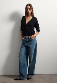 Schwarzer, tief ausgeschnittener Cardigan mit V-Ausschnitt, kombiniert mit lässigen blauen Jeans. Spitz zulaufende schwarze High Heels runden das Outfit ab. Weiche Textur, lässiges Design.