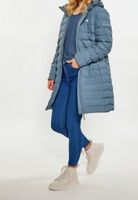 Vaaleansininen puhalla takki, jossa on quiltattu pinta, vetoketjukiinnitys ja irrotettava huppu, yhdistettynä tummansinisiin tiukkoihin farkkuihin ja vaaleisiin lenkkareihin.