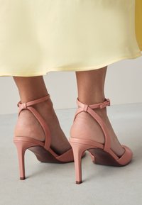Chaussures à talons hauts rose clair en cuir lisse, dotées de brides à la cheville et d'un talon effilé, posées sur un fond neutre.