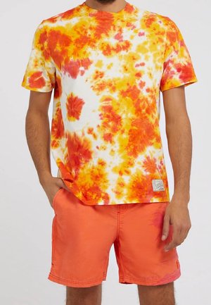 Mężczyzna w pomarańczowo-żółtej koszulce tie-dye i jaskrawo pomarańczowych szortach, stojący z jedną ręką w kieszeni, drugą swobodnie opartą.