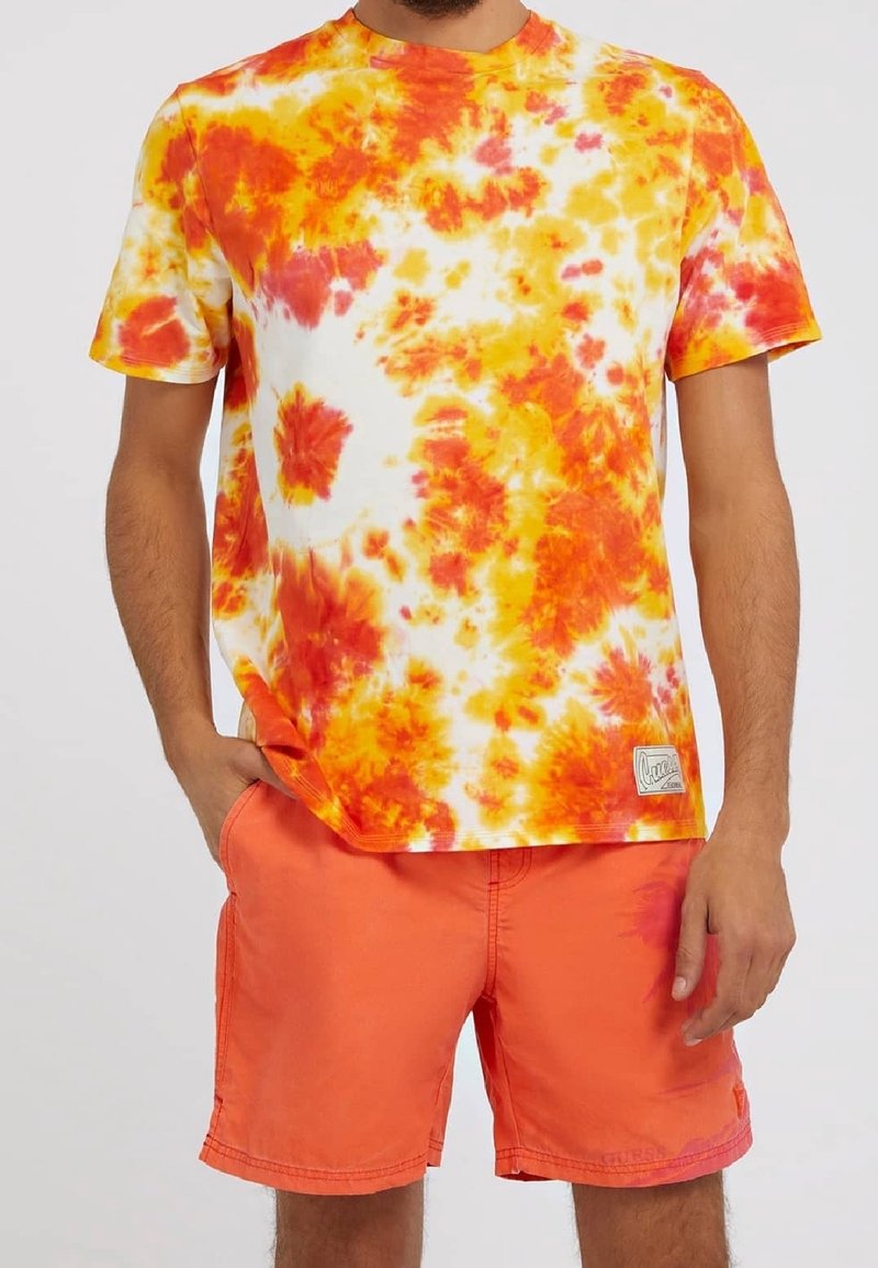 Homme portant un t-shirt tie-dye orange et jaune avec un short orange vif, debout avec une main dans la poche et l’autre détendue.