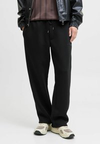 Schwarze Jogginghose aus weichem Material, mit einem Kordelzugbund, lockerer Passform und Seitentaschen. Kombiniert mit beigen Sportschuhen.