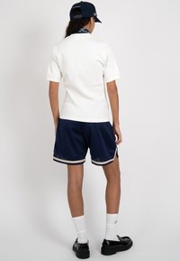 Personne portant une chemise blanche à manches courtes, un short en mesh bleu marine avec une bordure beige, des chaussettes blanches, des chaussures noires, une casquette bleu marine et un foulard bleu à motifs autour du cou.