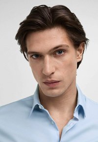 Chemise à col bleu clair avec une texture lisse, patte de boutonnage et finition douce, portée par un jeune homme aux cheveux bruns et aux yeux bleus.