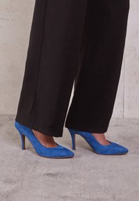 Blå mocka högklackade skor med spetstoppdesign. Kombinationen med vida svarta byxor, mot en neutral texturerad bakgrund.