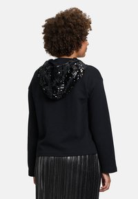 Felpa nera con cappuccio decorato da paillettes. Caratterizzata da una vestibilità comoda, maniche lunghe e orlo a coste. Indossata con una gonna plissè argentata.