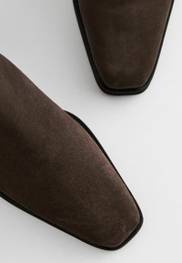 Hnědé semišové boty s vykrojenou špičkou a kontrastní černou podrážkou. Textura se zdá být měkká s matným povrchem.