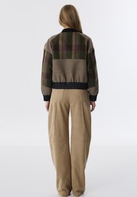 Wollen bomberjack in groen en bruin nadat, zwarte geribbelde kraag en boorden, gecombineerd met beige wijde pantalon met naaddetails.
