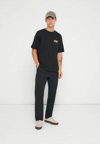 Zwart T-shirt met korte mouwen en gele grafiek, gecombineerd met donkere broeken en beige sneakers, afgemaakt met een lichtgekleurde pet.