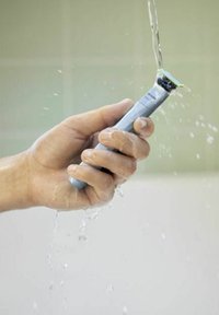 Elektrische scheerapparaat in een hand, grijze behuizing, groene en zwarte kop, water spat overal. Glad, ergonomisch ontwerp geschikt voor gebruik in de natte omstandigheden.