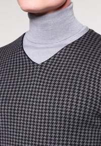 Col roulé gris superposé sous un gilet noir et gris à motif pied-de-poule avec un col en V et un tissu texturé.