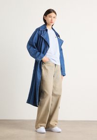 Duga traper jakna s velikim ovratnikom, slobodnog kroja, nosi se preko svijetloplave majice i bež širokih hlača; bijele tenisice upotpunjuju outfit.