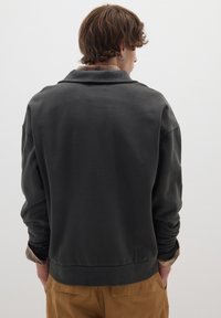 Felpa grigia scura con colletto e una cucitura texture visibile all'altezza del collo. Presenta una vestibilità comoda e polsini a costine, indossata sopra una camicia di flanella a quadri.