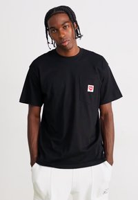 Black cotton t-shirt with a crew neckline, short sleeves, and a chest pocket featuring a red lips patch. Smooth texture and relaxed fit.
