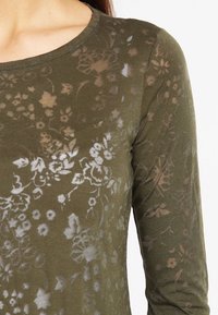 Olivegrön långärmad topp i mjukt, halvgenomskinligt tyg med ett blommönster i ljusare nyanser. Rund halsringning och figurnära design.