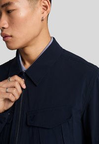 Giacca blu navy con colletto, chiusura con zip e due tasche frontali; realizzata in un tessuto liscio con dettagli cuciti.