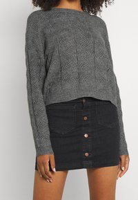 Pull en maille torsadée gris avec encolure ronde et coupe courte, associé à une jupe en denim noir agrémentée de boutons en bronze.