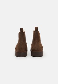 Botas de tobillo de ante marrón con una textura suave, punteras redondeadas y una suela de goma marrón baja. Presenta tiradores en la parte posterior para facilitar su puesta.