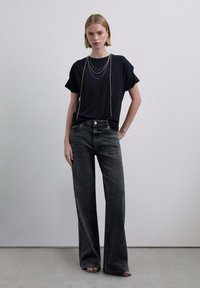 Camiseta negra de manga corta con tres cadenas plateadas en capas. Combinada con vaqueros grises de pierna ancha y talle alto. Texturas suaves, diseño minimalista.