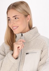 Giacca in pile di un beige chiaro con colletto alto, tessuto testurizzato, tasca sul petto con zip e maniche lisce. Materiale morbido e soffice molto evidente.