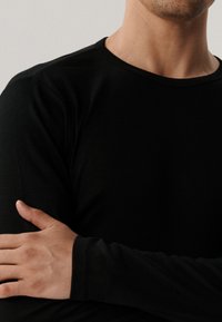 Czarna bluzka z długim rękawem wykonana z miękkiego materiału. Okrągły dekolt i dopasowany krój o gładkiej fakturze. Ręce skrzyżowane na ciele.